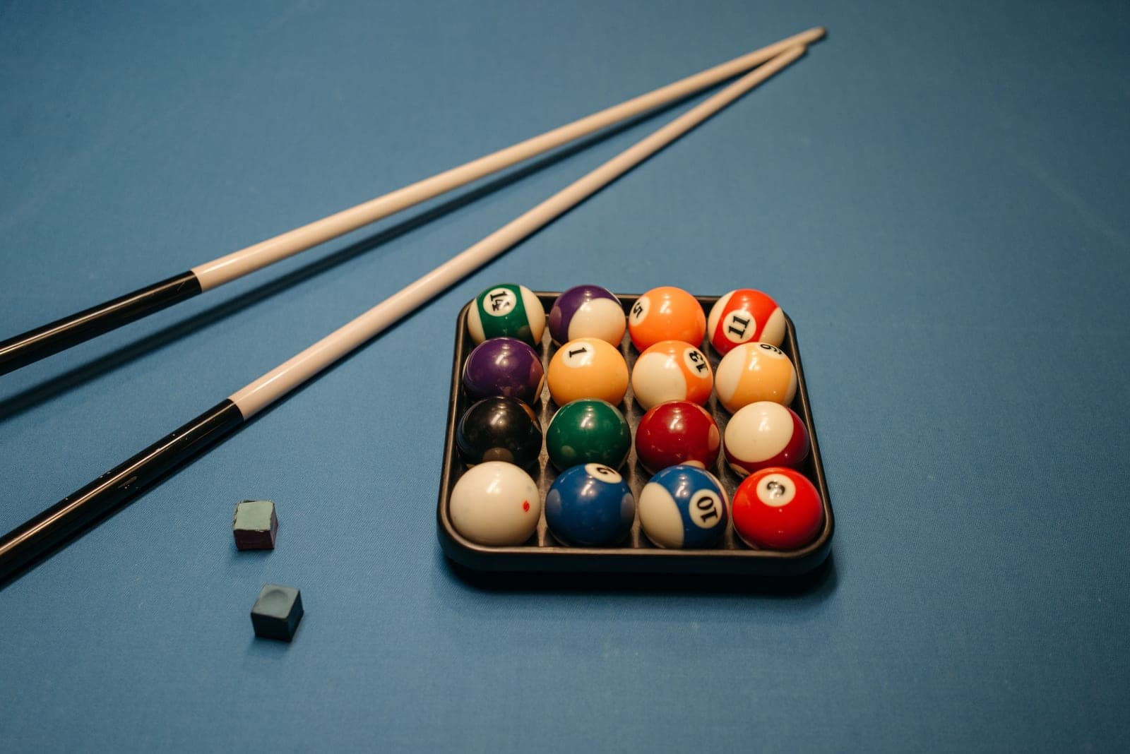 Pool cues racked on a wall beside chalk and a ball rack in a dark games room