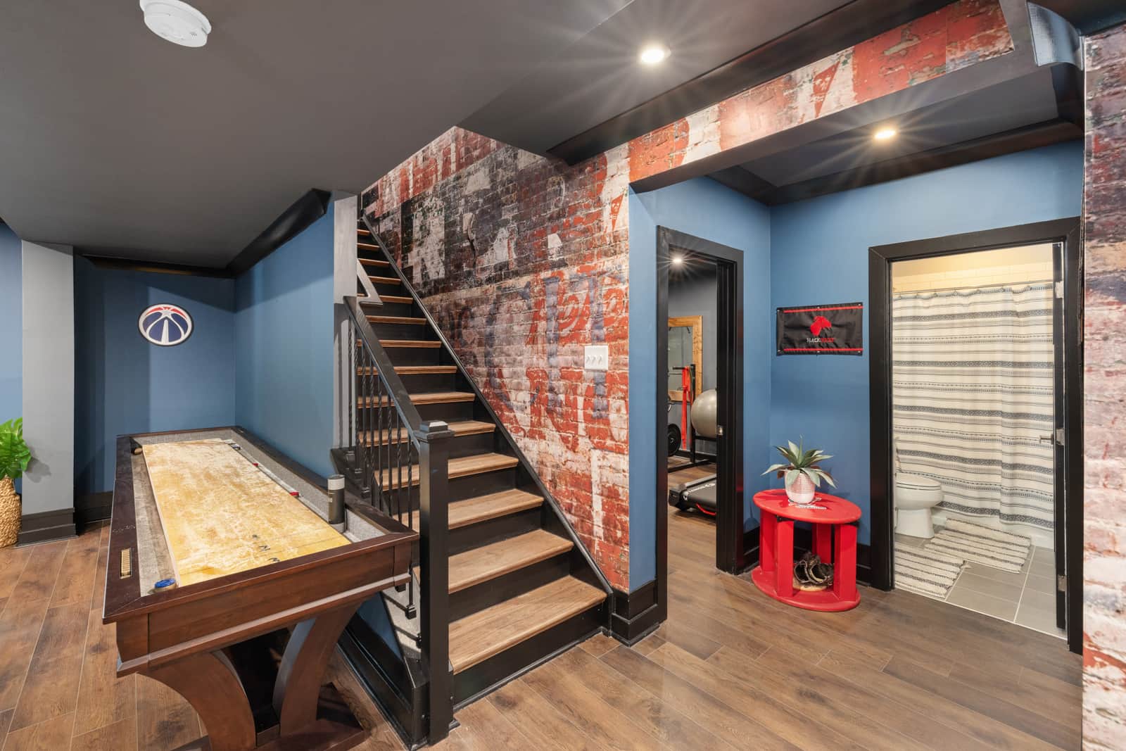 A wooden shuffleboard table along the wall of a finished basement, with the stairs beside it